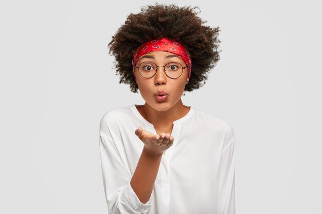 Hermosa tierna mujer negra con corte de pelo afro, sopla airkiss, mantiene la palma hacia la boca, expresa amor a distancia