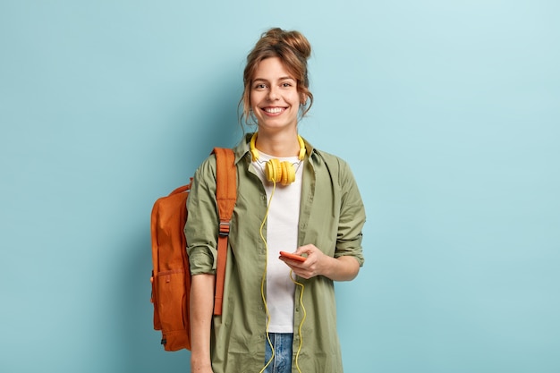 La hermosa viajera smilig tiene tiempo libre, disfruta de la comunicación en línea, está conectada a auriculares, escucha música de la lista de reproducción, usa una camiseta blanca informal y una camisa verde, lleva una mochila