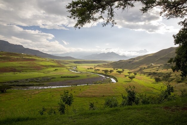 Hermosa vista del río, árboles y pastos rodeados de montañas con un cielo azul nublado