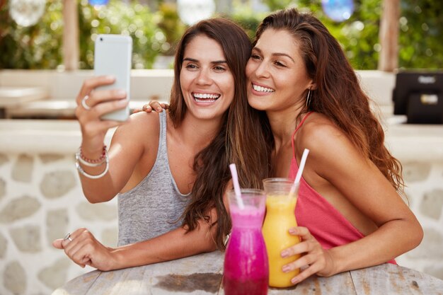 Hermosas amigas posan en la cámara del teléfono inteligente moderno, hacen selfie, se sientan juntas en un café al aire libre, beben batidos, tienen expresiones positivas.
