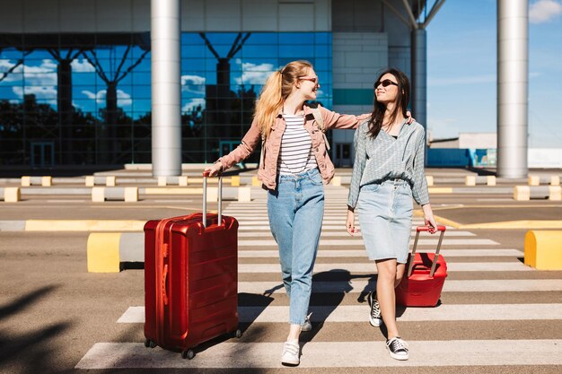 Hermosas chicas elegantes con gafas de sol caminando alegremente por la franja peatonal con maletas rojas y el aeropuerto en el fondo