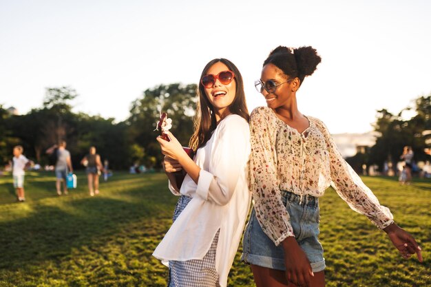 Hermosas chicas con gafas de sol jugando alegremente con una pequeña guitarra y bailando mientras pasan tiempo juntas en el parque