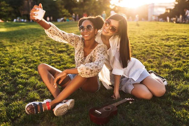 Hermosas chicas sonrientes sentadas en el césped felizmente tomando fotos en el celular mientras pasan tiempo juntas en el parque de la ciudad