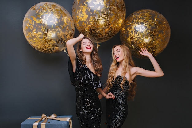 Hermosas damas bailando con las manos arriba frente a brillantes globos de helio y sonriendo. Foto interior de refinada cumpleañera de pelo castaño relajándose con un amigo y riendo.