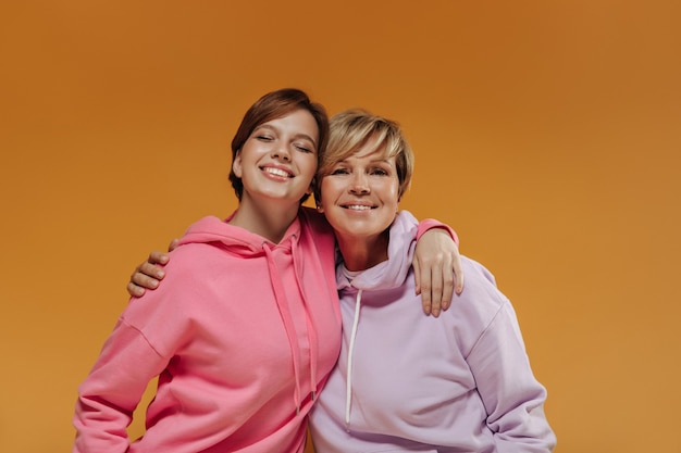 Hermosas dos damas con peinado corto y moderno en amplias sudaderas con capucha brillantes sonriendo y abrazándose sobre fondo naranja aislado.