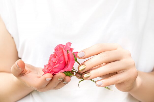 Hermosas manos femeninas con un diseño perfecto de uñas doradas y rosadas sostienen una rosa fresca