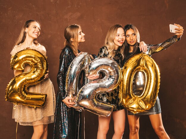 Hermosas mujeres celebrando el año nuevo. Felices y hermosas chicas con elegantes vestidos de fiesta sexys con globos dorados y plateados 2020, divirtiéndose en la fiesta de fin de año. Haciendo selfie o video para instagram