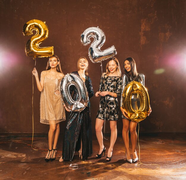 Hermosas mujeres celebrando el Año Nuevo. Felices y hermosas chicas con elegantes vestidos de fiesta sexys con globos dorados y plateados 2020, divirtiéndose en la fiesta de Nochevieja.