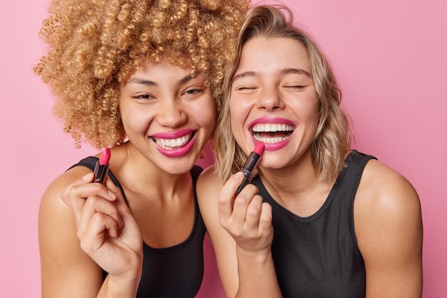 Hermosas mujeres felices se aplican pintalabios y se someten a procedimientos de belleza se paran una al lado de la otra vestidas con camisetas negras aisladas sobre fondo rosa Dos amigas se preparan para la fiesta quieren verse bien