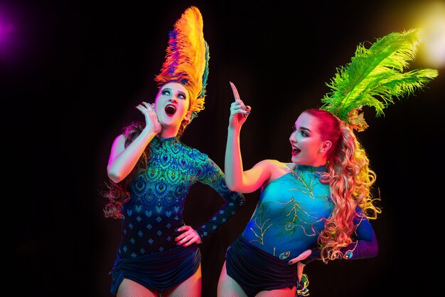 Hermosas mujeres jóvenes en carnaval, elegante disfraz de mascarada con plumas en pared negra en luz de neón