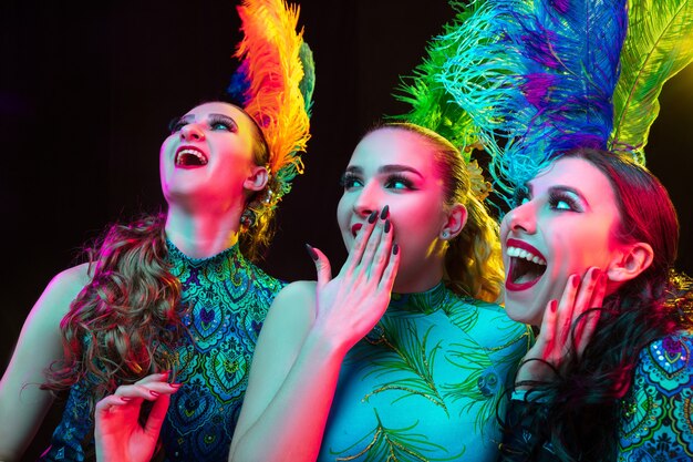 Hermosas mujeres jóvenes en carnaval, elegante disfraz de mascarada con plumas en pared negra en luz de neón