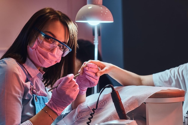 Foto gratuita el hermoso y alegre maestro de manicura está mirando a la cámara mientras trabaja en las uñas del cliente.