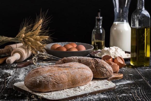 Foto gratuita hermoso bodegón con diferentes tipos de pan, cereales, harina en peso, espigas de trigo, jarra de leche y huevos sobre fondo negro