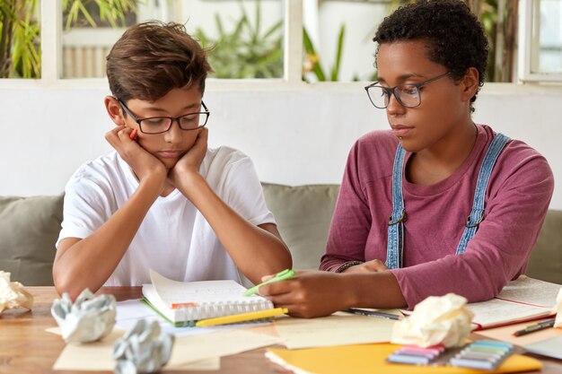 Hermoso chico asiático serio tiene entrenamiento de idiomas con un tutor, se sientan juntos en el escritorio, hacen los deberes y practican la lección, se enfocan en el bloc de notas con notas, se preparan para el examen o seminario universitario de ingreso