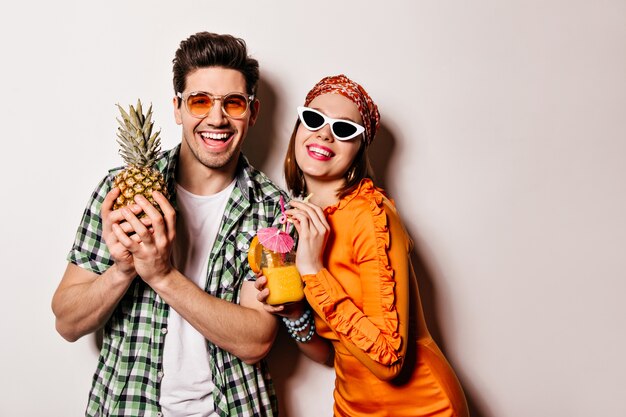 Hermoso chico y chica con gafas de sol y ropa de verano brillante están sonriendo y disfrutando de cócteles y piña.