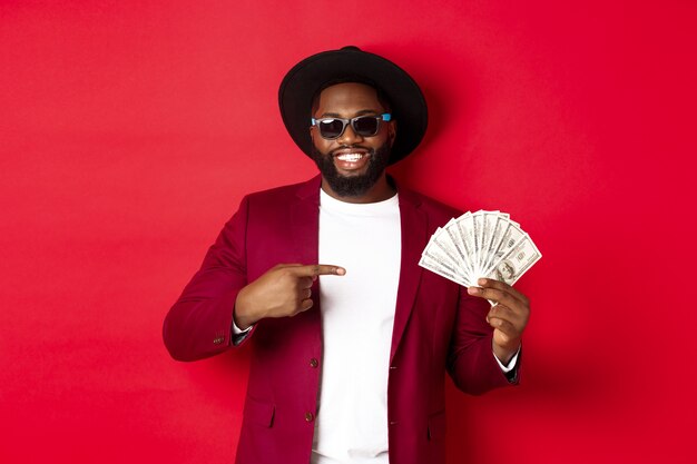 Hermoso y elegante modelo masculino afroamericano mostrando dinero y sonriendo, con gafas de sol y sombrero elegante, de pie sobre fondo rojo.