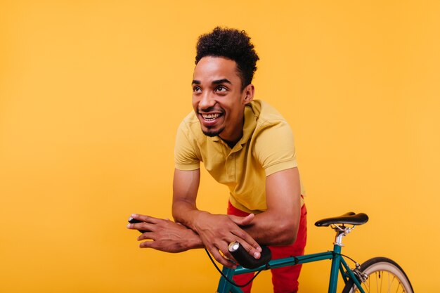 Hermoso hombre africano en camiseta riendo. Retrato de buen humor modelo masculino negro posando con bicicleta.