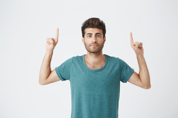 Hermoso hombre caucásico sin afeitar con expresión seria, en camiseta azul casual, apuntando con los dedos a la pared en blanco. Copiar espacio para publicidad.