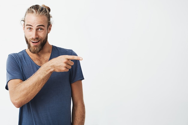 Hermoso hombre fuerte con peinado atractivo y barba en camisa azul apuntando a un lado con expresión de sorpresa