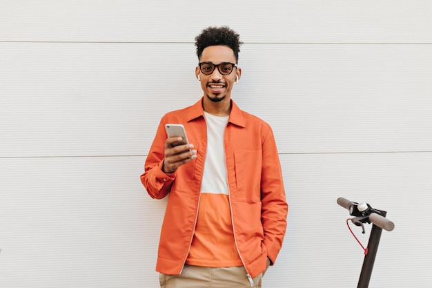 Hermoso hombre moreno rizado de piel oscura con gafas de sol de chaqueta naranja y pantalones cortos beige sonríe mira a la cámara sostiene el teléfono y posa cerca de la scooter