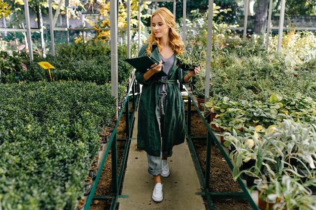 Hermoso jardín botánico, lleno de muchas flores y arbustos verdes. Chica con cabello rubio rizado, posa, presentándose como bióloga.