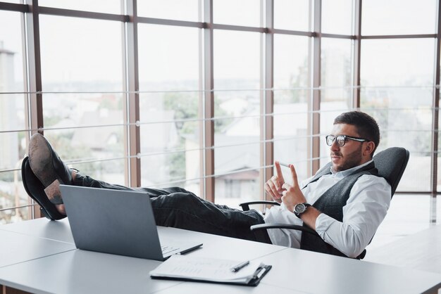 Hermoso joven empresario con gafas sosteniendo sus piernas sobre la mesa mirando una computadora portátil en la oficina