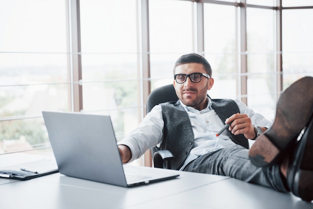 Hermoso joven empresario con gafas sosteniendo sus piernas sobre la mesa mirando una computadora portátil en la oficina