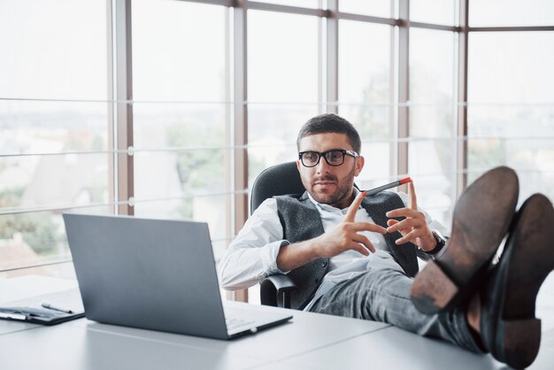 Hermoso joven empresario con gafas sosteniendo sus piernas sobre la mesa mirando una computadora portátil en la oficina