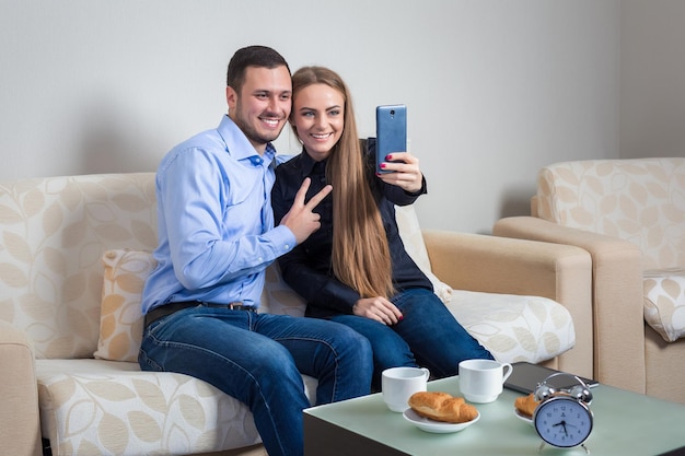 Hermoso joven y mujer haciendo selfie con cámara de teléfono, gente feliz tomando fotos sonriendo a la cámara, sentada en un sofá