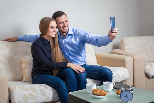 Hermoso joven y mujer haciendo selfie con cámara de teléfono, gente feliz tomando fotos sonriendo a la cámara, sentada en un sofá