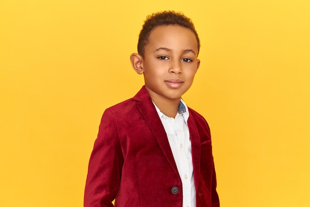 Hermoso lindo afroamericano poco posando aislado con expresión facial segura, sonriendo, vistiendo una chaqueta de terciopelo carmesí de moda.