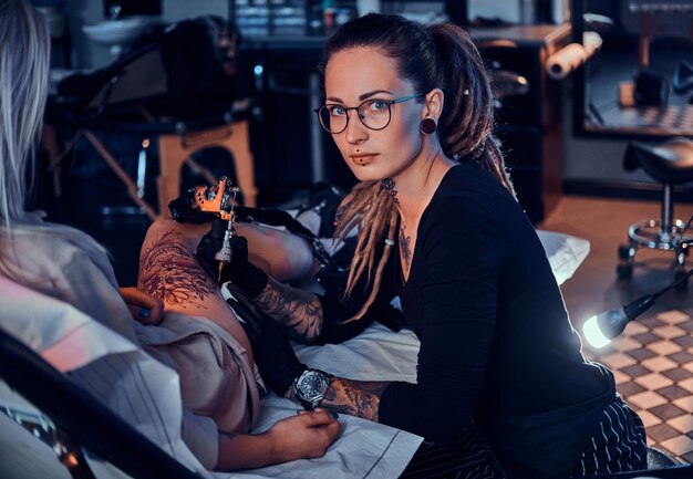 El hermoso maestro creativo con rastas está trabajando en un nuevo tatuaje de pierna grande para el cliente.