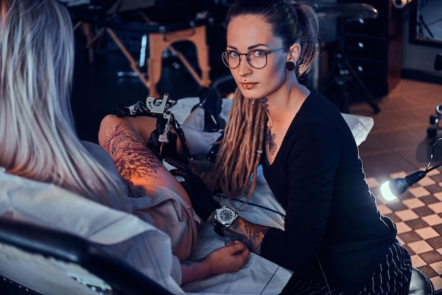 El hermoso maestro creativo con rastas está trabajando en un nuevo tatuaje de pierna grande para el cliente.