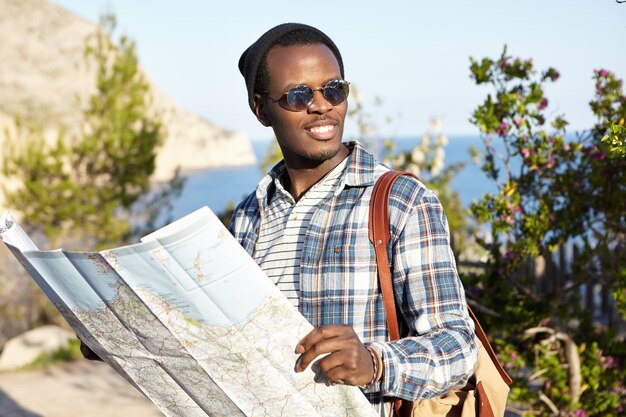 Hermoso mochilero negro de moda con guía de viaje de papel en sus manos buscando restaurante vegetariano mientras explora la ciudad europea durante el viaje