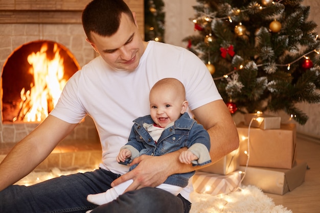 Hermoso padre brunet sosteniendo a su pequeña hija en las manos, mirando a su lindo y encantador niño, sentado junto a la chimenea y el árbol de Navidad en casa, feliz año nuevo.