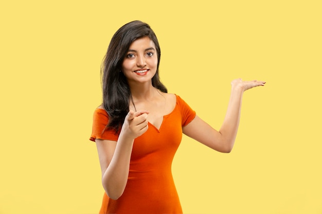 Hermoso retrato femenino de medio cuerpo aislado en espacio amarillo. Joven mujer india emocional en vestido apuntando y mostrando. Espacio negativo