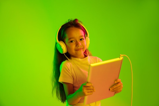 Hermoso retrato femenino de medio cuerpo aislado sobre fondo verde en luz de neón. Chica joven emocional. Las emociones humanas, el concepto de expresión facial. Colores de moda. Usando tableta para juegos, vlog, selfie.