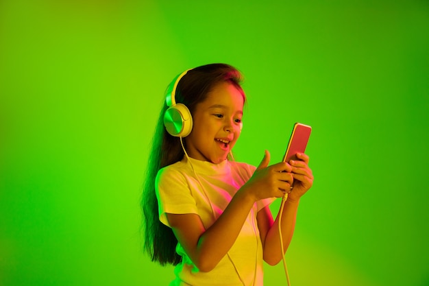 Hermoso retrato femenino de medio cuerpo aislado sobre fondo verde en luz de neón. Chica joven emocional. Las emociones humanas, el concepto de expresión facial. Uso de teléfonos inteligentes para vlog, selfies, chats, juegos.