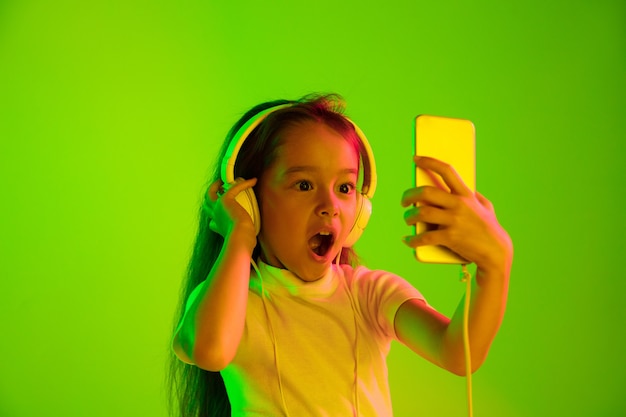 Hermoso retrato femenino de medio cuerpo aislado sobre fondo verde en luz de neón. Chica joven emocional. Las emociones humanas, el concepto de expresión facial. Uso de teléfonos inteligentes para vlog, selfies, chats, juegos.