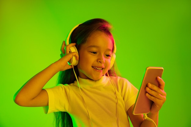 Hermoso retrato femenino de medio cuerpo aislado sobre fondo verde en luz de neón. Chica joven emocional. Las emociones humanas, el concepto de expresión facial. Uso de teléfonos inteligentes para vlog, selfies, chats, juegos.