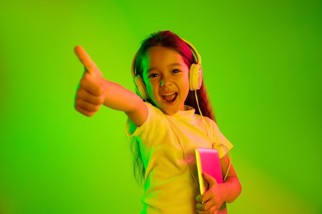 Hermoso retrato femenino de medio cuerpo aislado sobre fondo verde en luz de neón. Joven adolescente emocional. Las emociones humanas, el concepto de expresión facial. Colores de moda. Sosteniendo la tableta y sonriendo.