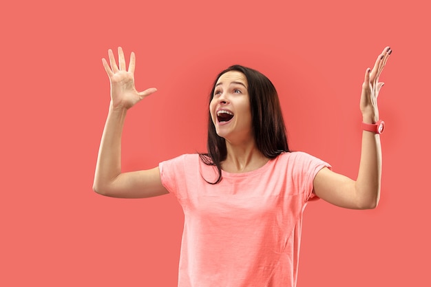 Hermoso retrato frontal de medio cuerpo femenino aislado sobre fondo de estudio coral. Joven mujer sorprendida emocional de pie con la boca abierta.