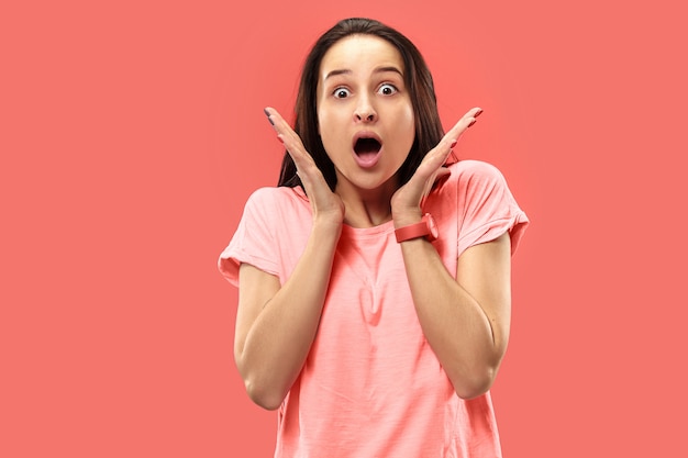 Hermoso retrato frontal de medio cuerpo femenino aislado sobre fondo de estudio coral. Joven mujer sorprendida emocional de pie con la boca abierta.