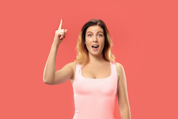 Hermoso retrato frontal de medio cuerpo femenino aislado sobre fondo de estudio coral. Joven mujer sorprendida emocional de pie con la boca abierta.