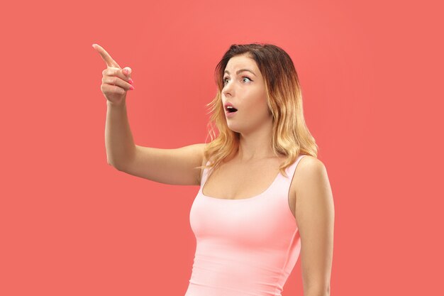 Hermoso retrato frontal de medio cuerpo femenino aislado sobre fondo de estudio coral. Joven mujer sorprendida emocional de pie con la boca abierta.