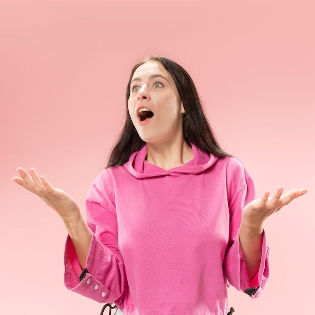 Hermoso retrato frontal de medio cuerpo femenino aislado sobre fondo rosa de estudio. Joven mujer sorprendida emocional de pie con la boca abierta.