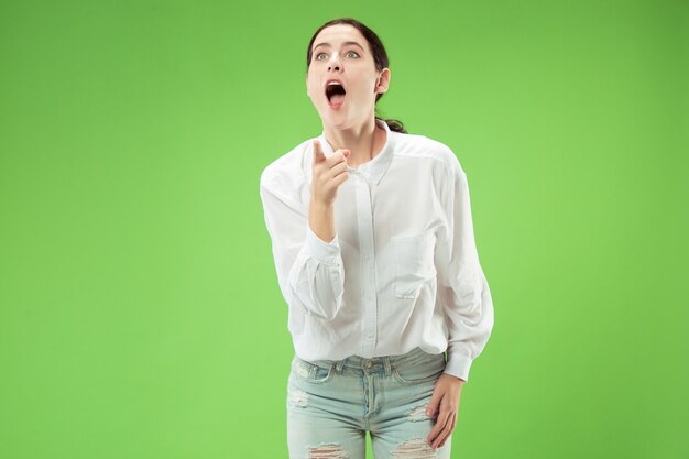 Hermoso retrato frontal de medio cuerpo femenino aislado sobre fondo verde de estudio. Joven mujer sorprendida emocional de pie con la boca abierta.