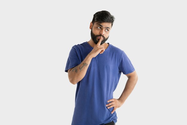 Hermoso retrato masculino de medio cuerpo aislado en la pared blanca. Joven hindú emocional en camisa azul. Expresión facial, emociones humanas, concepto publicitario. Pensando, susurrando un secreto.