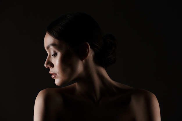 Hermoso retrato de mujer elegante semidesnuda con cabello oscuro en moño poniendo la cabeza a un lado, aislado sobre negro