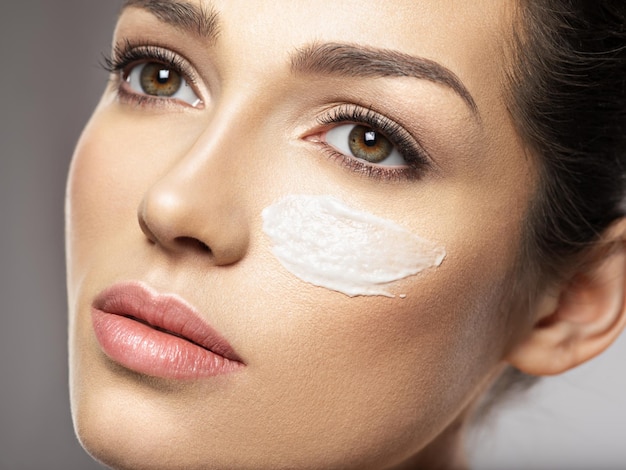 Hermoso rostro de mujer joven con frotis de crema cosmética en la cara cerca del ojo. Concepto de cuidado de la piel. Concepto de tratamiento de belleza.